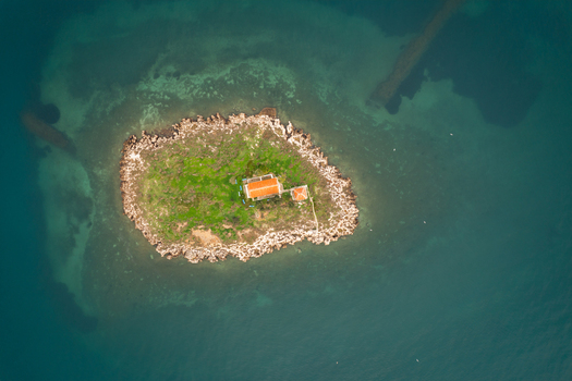 Aerial view of a small island featuring a church with a terracotta roof surrounded by turquoise sea water and rocky shores in Itea Greece.