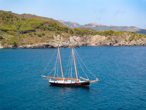 Aerial view of a classic two masted sailboat cruising on the deep blue waters of the Mediterranean Sea near the rocky coastline and green hills of Lacona Tuscany Italy.