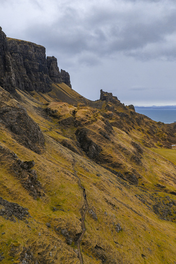 Aerial view of The Quiraing s dramatic rocky cliffs and steep grassy slopes with hikers on a narrow trail overlooking the sea in Isle of Skye Scotland United Kingdom. Aerial view of The Quiraing s dramatic rocky cliffs and steep grassy slopes with hikers on a narrow trail overlooking the sea in Isle of Skye Scotland United Kingdom.