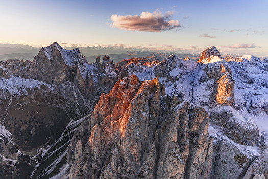 Aerial view of jagged snow dusted Dolomites Catinaccio Rosengarten peaks blaze with golden light against the cool blue sky Trentino South Tyrol Italy.