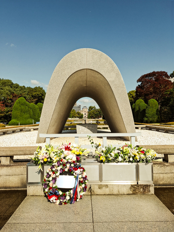 View of the Hiroshima Peace Memorial Cenotaph with floral wreaths and the Atomic Bomb Dome in the background under a clear blue sky in Hiroshima Hiroshima Japan. View of the Hiroshima Peace Memorial Cenotaph with floral wreaths and the Atomic Bomb Dome in the background under a clear blue sky in Hiroshima Hiroshima Japan.