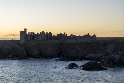 Aerial view of the silhouette of ruined Slains Castle on rugged cliffs overlooking the North Sea during a golden sunset Cruden Bay Scotland United Kingdom. Aerial view of the silhouette of ruined Slains Castle on rugged cliffs overlooking the North Sea during a golden sunset Cruden Bay Scotland United Kingdom.