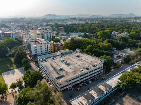 Aerial view of a multi story parking structure with cars parked on the rooftop surrounded by dense urban greenery and residential buildings in Udaipur Rajasthan India.