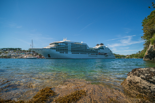 View of the Seabourn Ovation cruise ship anchored in the Fowey River with the historic town of Fowey in the background under a clear blue sky in Fowey Cornwall United Kingdom. View of the Seabourn Ovation cruise ship anchored in the Fowey River with the historic town of Fowey in the background under a clear blue sky in Fowey Cornwall United Kingdom.