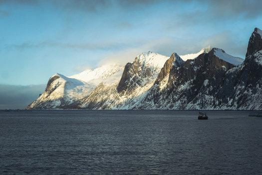 View of sunlight kisses the snow capped peaks rising majestically from the dark tranquil waters a lone boat sailing into the distance Indregård Troms Norway. View of sunlight kisses the snow capped peaks rising majestically from the dark tranquil waters a lone boat sailing into the distance Indregård Troms Norway.