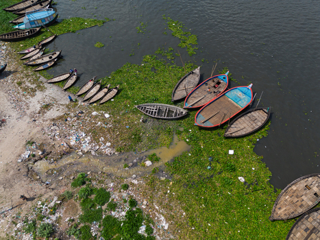 Aerial view of the Buriganga River with traditional wooden boats and people on the shore near green water hyacinth in Dhaka Dhaka Division Bangladesh.