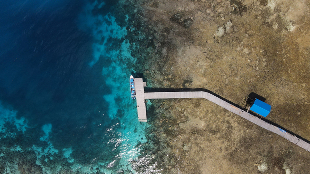 Aerial view of a wooden pier extending into the turquoise waters of Raja Ampat with a small boat docked and a blue roofed shelter Raja Ampat Southwest Papua Indonesia.