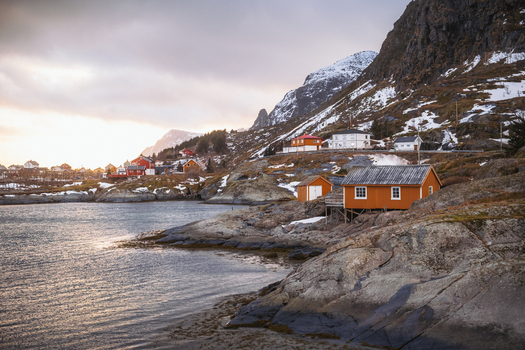 View of rustic red houses nestled on rocky shores under snow dusted mountains where the sea meets the sky in Tind Nordland Norway.