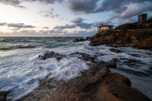 View of the rocky coastline and the historic tower of Calafuria with crashing waves and a dramatic cloudy sky at sunset Calafuria Toscana Italy.