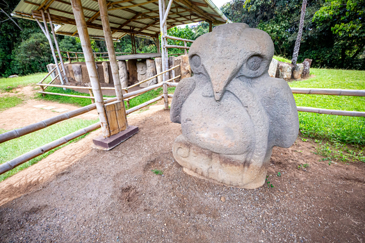 View of a pre Columbian stone statue of a bird like figure at San Agustin Archaeological Park with a bamboo shelter structure in the background San Agustin Huila Colombia.