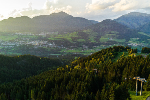 Aerial view of Fiemme Valley with dense pine forests rolling mountain slopes and a small village in the distance under a warm sunset sky in Trentino Alto Adige Italy.
