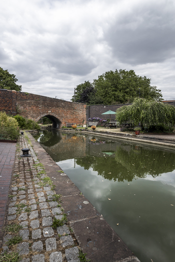 View of the historic brick bridge over the Coventry Canal with a cobblestone path lush greenery and a picnic area in Coventry England United Kingdom.