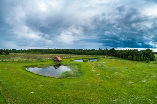 Aerial view of a small wooden house with a red roof situated between two tranquil ponds in a lush green meadow surrounded by dense forest under a dramatic stormy sky in Latvia. Aerial view of a small wooden house with a red roof situated between two tranquil ponds in a lush green meadow surrounded by dense forest under a dramatic stormy sky in Latvia.