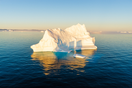 Aerial view of a large iceberg floating in the deep blue ocean illuminated by the warm golden light of the setting sun with reflections on the water surface in Greenland. Aerial view of a large iceberg floating in the deep blue ocean illuminated by the warm golden light of the setting sun with reflections on the water surface in Greenland.