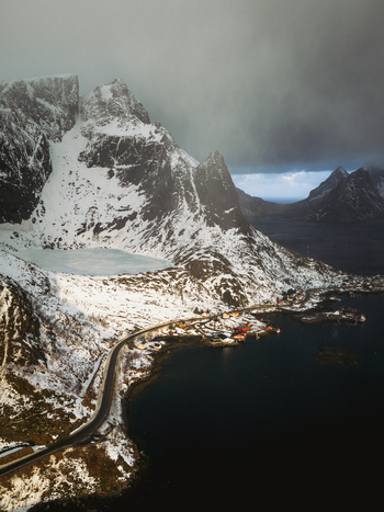 Aerial view of snow dusted peaks cascade into icy fjords a winding road hugs the coastline under a brooding sky Reinebringen Nordland Norway.