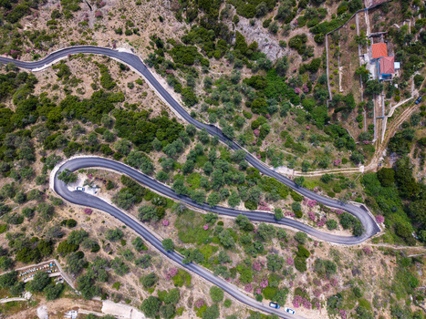 Aerial view of a winding mountain road with sharp hairpin turns snaking through a lush green hillside with olive trees and pink flowers in Qeparo Fshat Vlore County Albania. Aerial view of a winding mountain road with sharp hairpin turns snaking through a lush green hillside with olive trees and pink flowers in Qeparo Fshat Vlore County Albania.