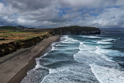 Aerial view of Praia do Areal de Santa Barbara with its dark sand rolling ocean waves and green coastal fields under a cloudy sky in Ribeira Grande Acores Portugal. Aerial view of Praia do Areal de Santa Barbara with its dark sand rolling ocean waves and green coastal fields under a cloudy sky in Ribeira Grande Acores Portugal.