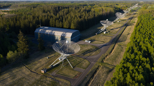 Aerial view of the Westerbork Synthesis Radio Telescope dishes lined up along a forest path with a large hangar building under clear sunlight in Westerbork Drenthe Netherlands. Aerial view of the Westerbork Synthesis Radio Telescope dishes lined up along a forest path with a large hangar building under clear sunlight in Westerbork Drenthe Netherlands.