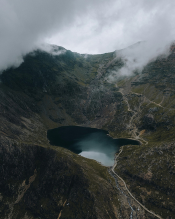 Aerial view of the rugged slopes of Snowdon surrounding a dark mountain lake with a winding hiking path under a cloudy sky in Caernarfon Wales United Kingdom.