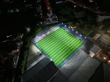 Aerial view of Bravo Arena soccer field illuminated at night with artificial lights surrounded by urban buildings and parked cars in Kecamatan Denpasar Selatan Bali Indonesia.