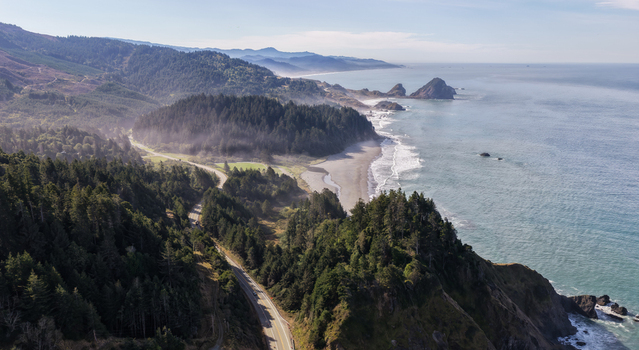 Aerial view of Highway 101 winding through dense evergreen forests along the rugged coastline with rocky cliffs and sandy beaches in Port Orford Oregon United States. Aerial view of Highway 101 winding through dense evergreen forests along the rugged coastline with rocky cliffs and sandy beaches in Port Orford Oregon United States.