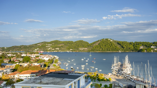 View of the harbor in Lefkada with numerous sailboats moored at the docks surrounded by lush green hills and clear blue sky in Lefkada Greece.