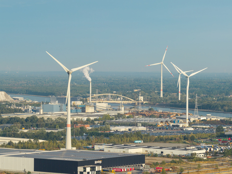 Genk Belgium 27 August 2026 Aerial view of wind turbines in an industrial landscape with warehouses a canal and a bridge under a clear blue sky.