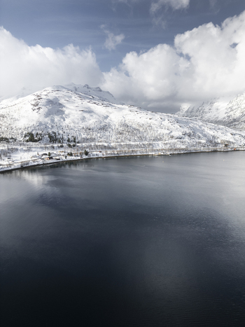 Aerial view of the snow covered mountains and the dark waters of Grotfjord with a small coastal settlement along the shore in Tromvik Troms Norway. Aerial view of the snow covered mountains and the dark waters of Grotfjord with a small coastal settlement along the shore in Tromvik Troms Norway.