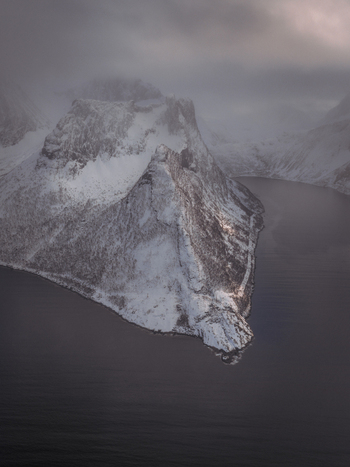 Aerial view of snow dusted peaks pierce through the misty veil meeting the calm dark waters below Fjordgård Troms Norway. Aerial view of snow dusted peaks pierce through the misty veil meeting the calm dark waters below Fjordgård Troms Norway.