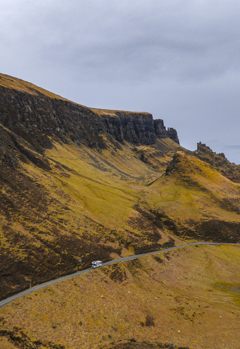Aerial view of The Quiraing landscape with a white campervan driving on a winding road under an overcast sky in Isle of Skye Scotland United Kingdom. Aerial view of The Quiraing landscape with a white campervan driving on a winding road under an overcast sky in Isle of Skye Scotland United Kingdom.