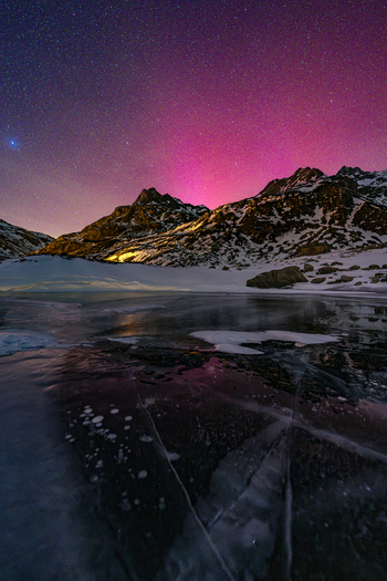 View of a frozen lake with ice bubbles and snow covered mountains under a vibrant pink aurora borealis and starry night sky in Madesimo Lombardy Italy. View of a frozen lake with ice bubbles and snow covered mountains under a vibrant pink aurora borealis and starry night sky in Madesimo Lombardy Italy.