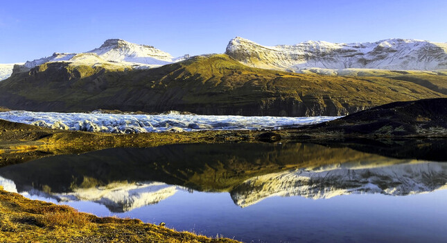 Aerial view of the Svinafellsjokull glacier with its icy blue tongue reflecting in a calm glacial lake surrounded by snow dusted mountains and rugged volcanic terrain in Svinafellsjokull Iceland.