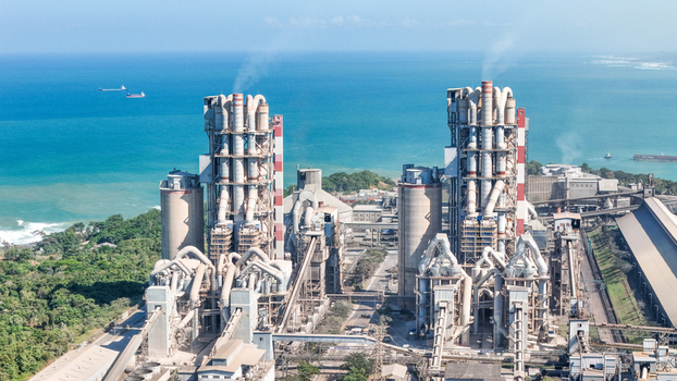 Aerial view of a large industrial cement factory complex with tall silos and processing towers situated along a lush green coastline overlooking the blue sea in Indonesia.