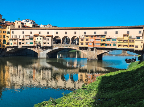 Firenze Italy 26 April 2026 View of the historic Ponte Vecchio bridge spanning the Arno river with medieval buildings and clear reflections in the water under a bright blue sky.