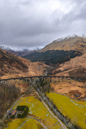 Aerial view of the historic Glenfinnan Viaduct with its stone arches spanning a rugged valley under snow capped mountains in Glenfinnan Scotland United Kingdom.