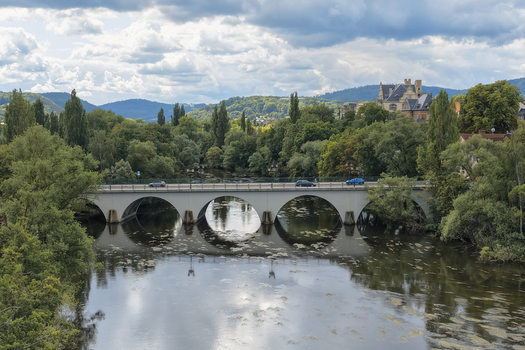 Aerial view of a multi arch stone bridge spanning the Saale River in Saalfeld surrounded by lush green trees and reflecting clouds in the water Saalfeld Thuringia Germany. Aerial view of a multi arch stone bridge spanning the Saale River in Saalfeld surrounded by lush green trees and reflecting clouds in the water Saalfeld Thuringia Germany.