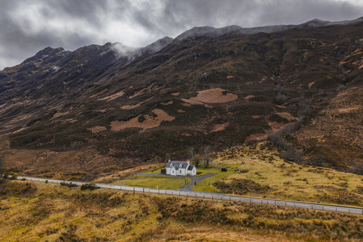 Aerial view of a remote white cottage at the base of rugged mountains under a dramatic cloudy sky Scotland United Kingdom. Aerial view of a remote white cottage at the base of rugged mountains under a dramatic cloudy sky Scotland United Kingdom.