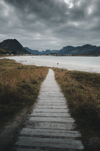 View of a wooden boardwalk leading to a sandy beach with distant mountains under a dramatic cloudy sky in Ramberg Nordland Norway. View of a wooden boardwalk leading to a sandy beach with distant mountains under a dramatic cloudy sky in Ramberg Nordland Norway.