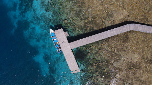 Aerial view of a boat docked at a wooden pier over vibrant coral reefs in the clear turquoise waters of Raja Ampat Southwest Papua Indonesia.