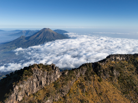 Aerial view of Gunung Sumbing Wonosobo rising above a sea of clouds with rugged rocky ridges in the foreground under a clear blue sky in Kecamatan Kalikajar Jawa Tengah Indonesia.