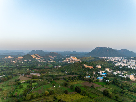 Aerial view of the historic City Palace complex nestled among the Aravalli hills with lush green fields and urban development in Udaipur Rajasthan India.