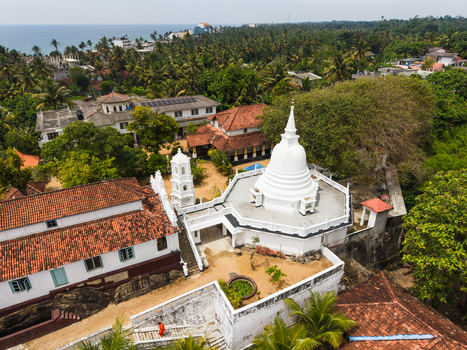 Aerial view of the white stupa of Kumarakanda Kumara Maha Viharaya surrounded by red tiled roofs and lush palm trees near the coast in Hikkaduwa Southern Province Sri Lanka. Aerial view of the white stupa of Kumarakanda Kumara Maha Viharaya surrounded by red tiled roofs and lush palm trees near the coast in Hikkaduwa Southern Province Sri Lanka.