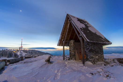 View of a small snow covered stone building with a wooden roof against a blue sky and a distant communication tower Schöckl Styria Austria. View of a small snow covered stone building with a wooden roof against a blue sky and a distant communication tower Schöckl Styria Austria.
