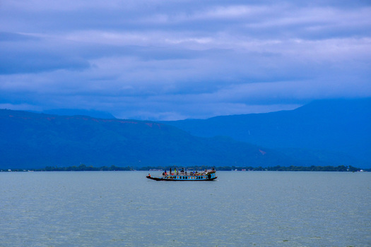 View of a boat sailing on the calm waters of Tanguar Haor with distant mountains under a cloudy sky in Sunamganj District Sylhet Division Bangladesh.