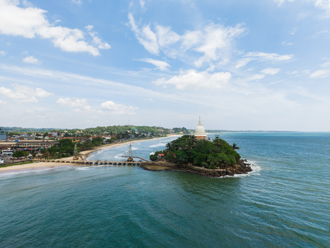 Aerial view of Parevi Duwa Temple on a small island connected by a bridge to the sandy beach and coastal town under a blue sky Matara Southern Province Sri Lanka. Aerial view of Parevi Duwa Temple on a small island connected by a bridge to the sandy beach and coastal town under a blue sky Matara Southern Province Sri Lanka.
