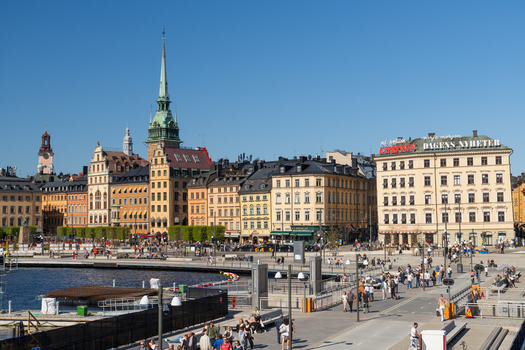 Slussen Sweden 02 May 2026 View of Vattentorget Slussen with people walking along the waterfront against the historic skyline of Gamla Stan under a clear blue sky.