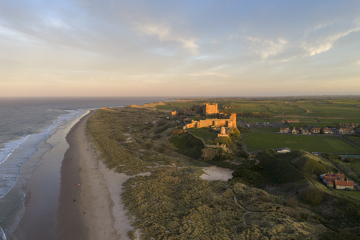 Aerial view of Bamburgh Castle overlooking the sandy beach and North Sea coastline during golden hour in Bamburgh England United Kingdom. Aerial view of Bamburgh Castle overlooking the sandy beach and North Sea coastline during golden hour in Bamburgh England United Kingdom.