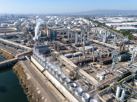 Aerial view of a large oil refinery complex featuring numerous storage tanks cooling towers emitting steam and complex piping networks in Carson California United States.