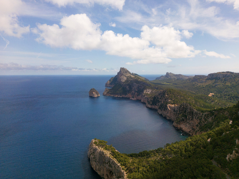 Aerial view of Cap de Formentor featuring dramatic limestone cliffs lush green vegetation and the deep blue Mediterranean Sea under a cloudy sky Balearic Islands Spain.