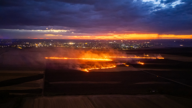 Aerial view of a field fire burning at night with glowing flames and smoke across dark agricultural land with the distant city lights of Sremska Mitrovica Vojvodina Serbia.